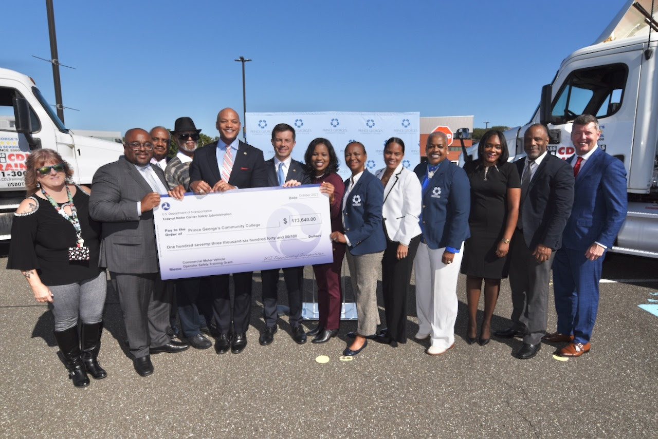 Governor Moore, U.S. Secretary of Transportation Pete Buttigieg, and others stand with a large check at Prince George's Community College.